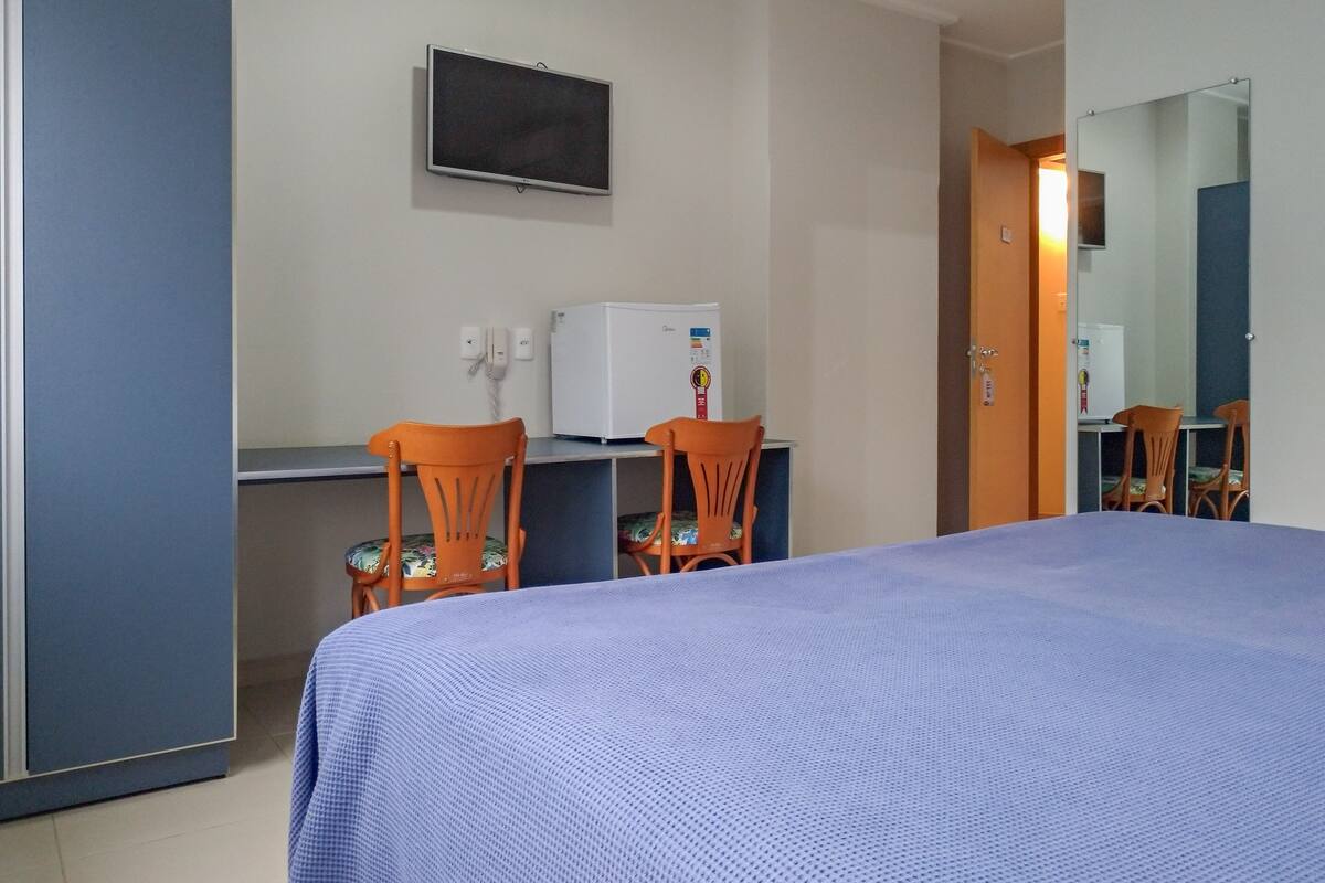 Quarto de hotel com cama, escrivaninha com computador, duas cadeiras, televisão e frigorífico.