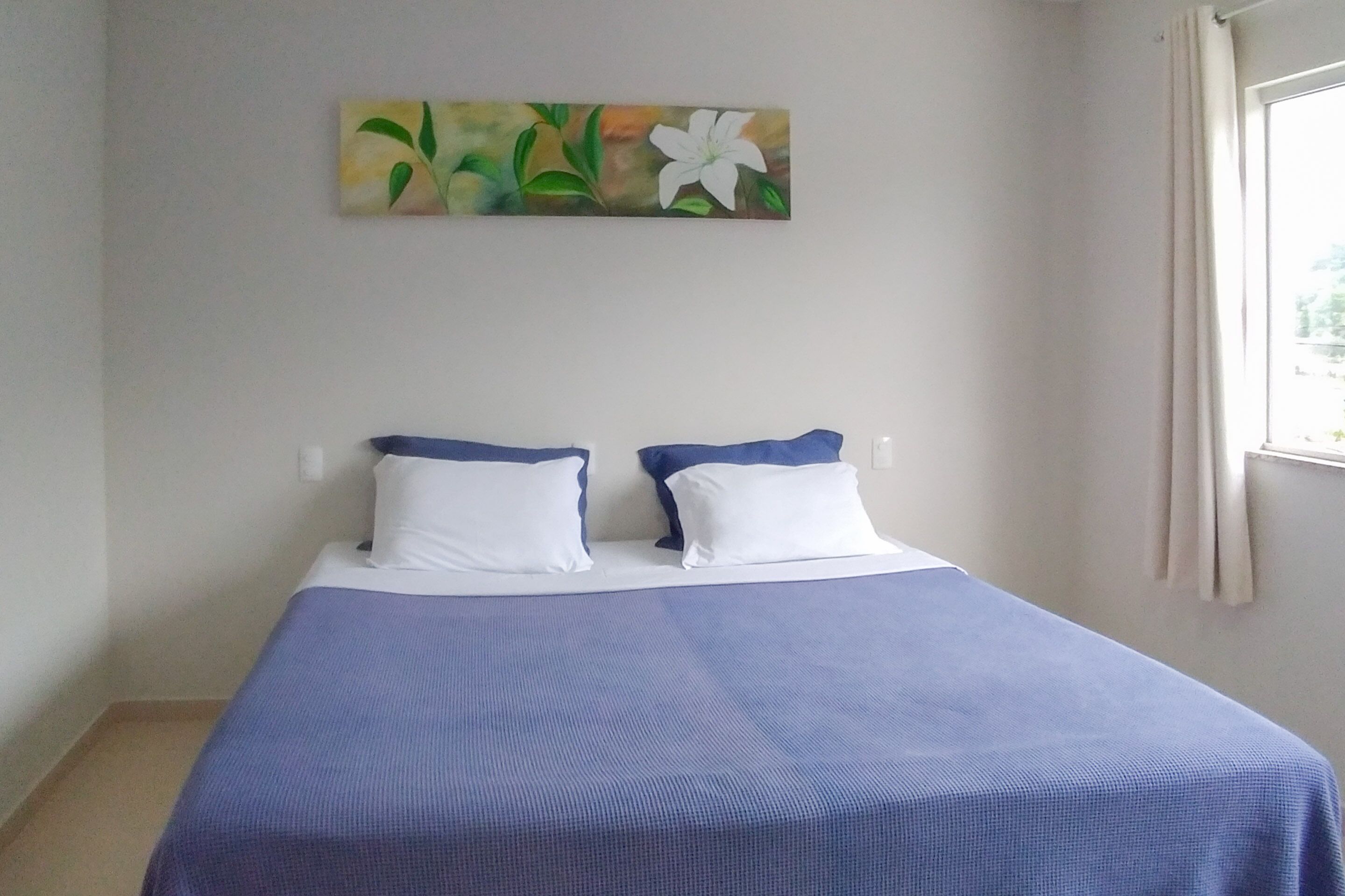 Una cama individual con una colcha azul y dos almohadas blancas, un cuadro de flores en la pared y una ventana con cortinas.