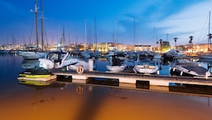 Ein Hafen im Abendlicht mit mehreren vertäuten Booten, einer Holzsteganlage und einer Stadtkulisse im Hintergrund.