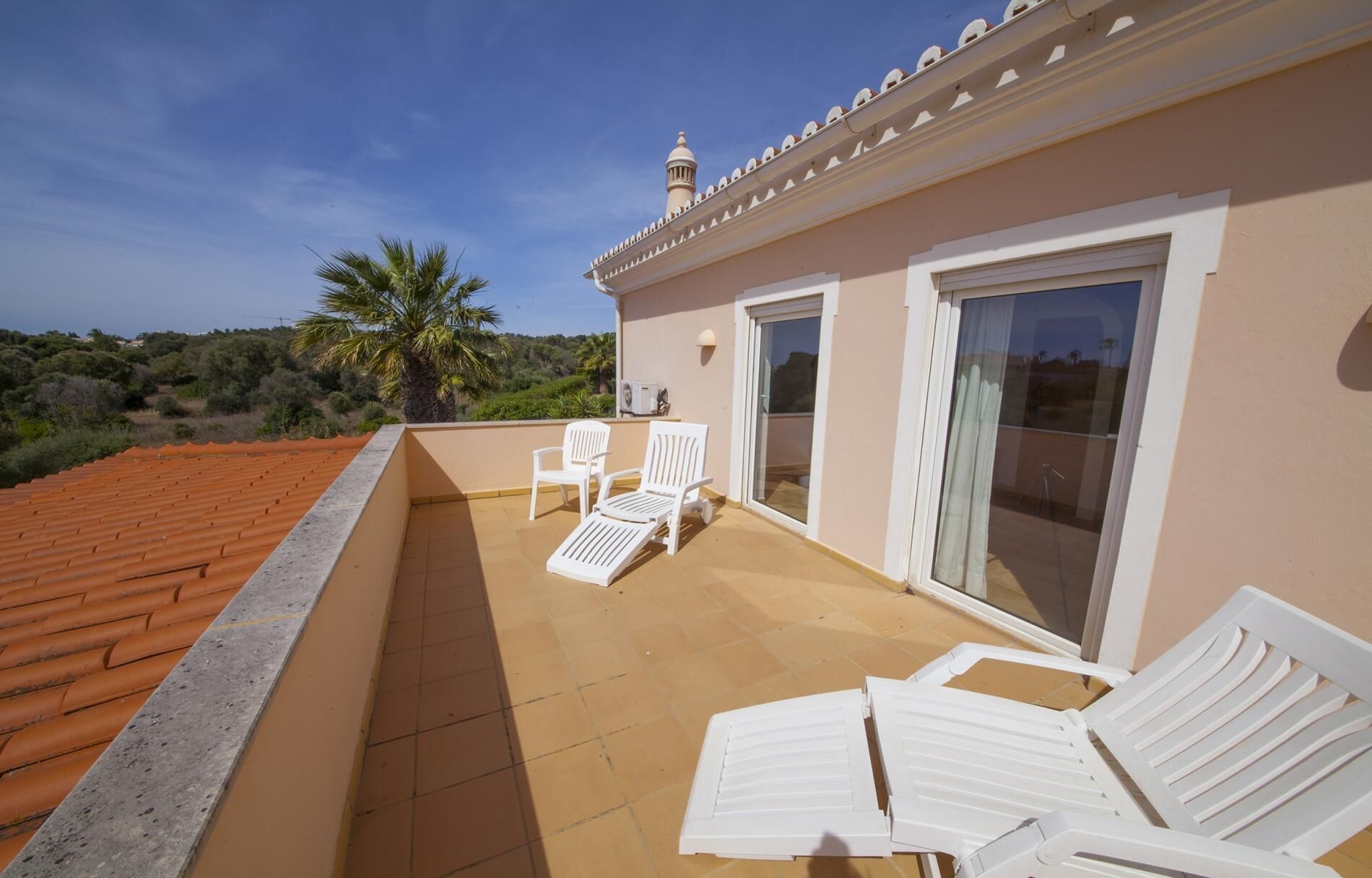 Eine Dachterrasse mit Fliesenboden, weißen Liegestühlen und einer Glastür zum Außenbereich.