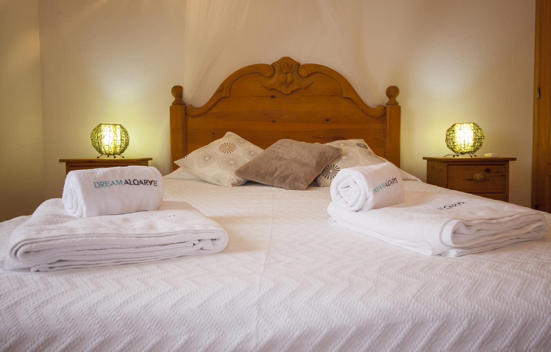 A neatly made bed with white linens and two towels with "Dream Algarve" text, a wooden headboard, and two bedside lamps.