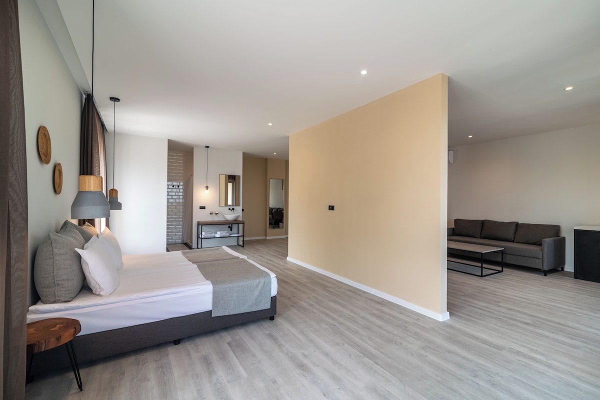 Un dormitorio moderno con cama, mesita de noche y lámpara. Cuenta con una sala de estar con sofá y mesa de centro. El cuarto tiene piso de madera y paredes color beige.