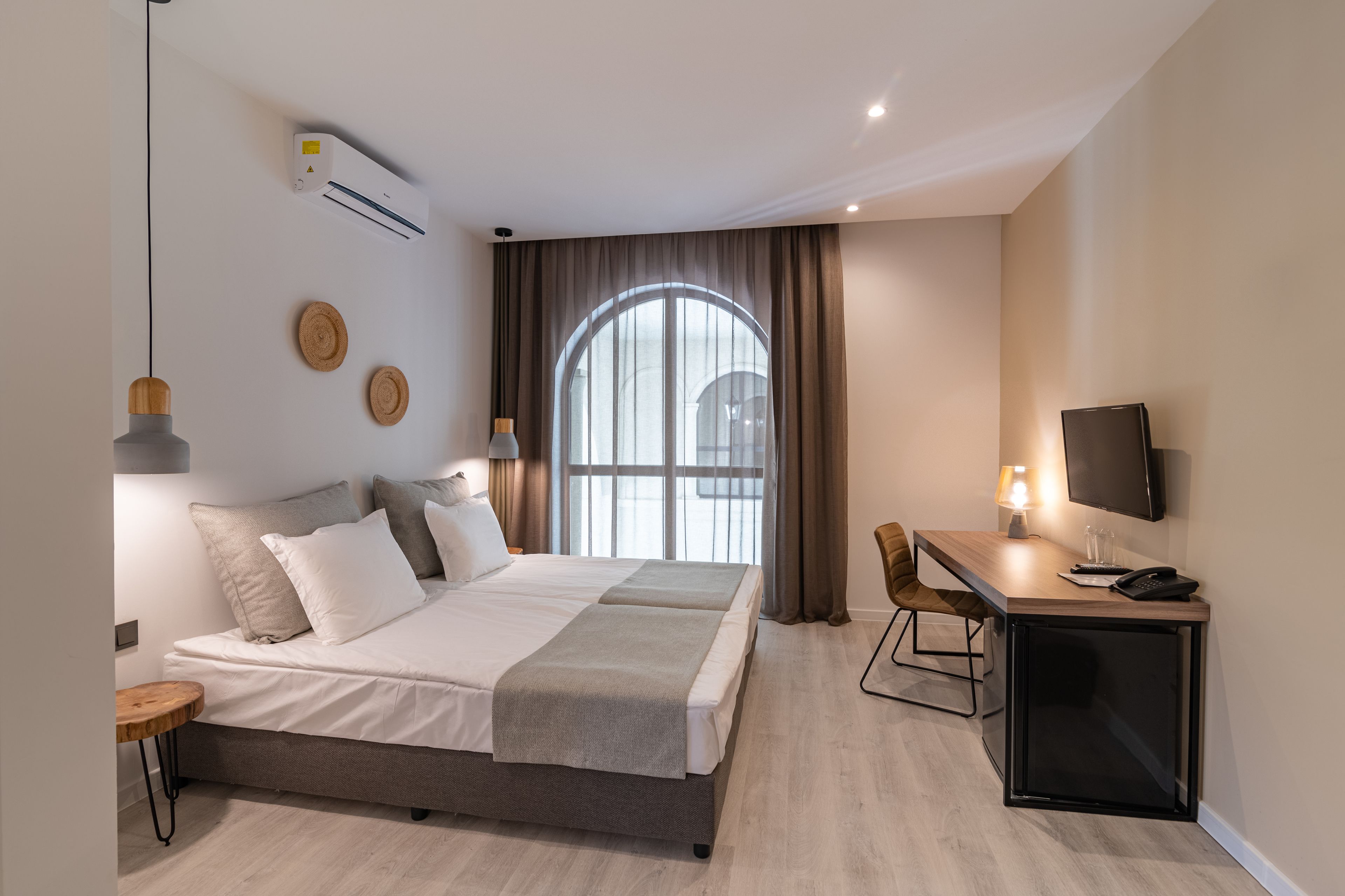 Moderni hotellihuone, jossa on suuri sänky, työpöytä tietokoneella, televisio ja ilmastointilaite.