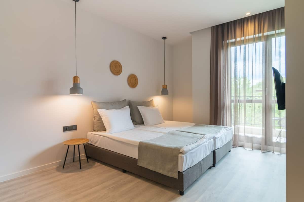 A modern bedroom with a double bed, two pendant lights, a small wooden stool, and a large window with curtains.