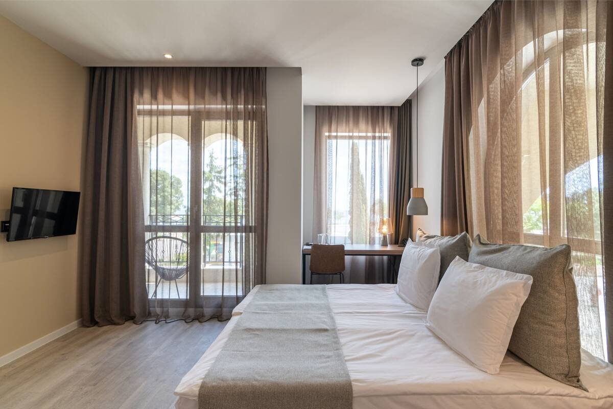 A hotel room with a bed, a desk, a chair, a TV, and large windows with curtains.