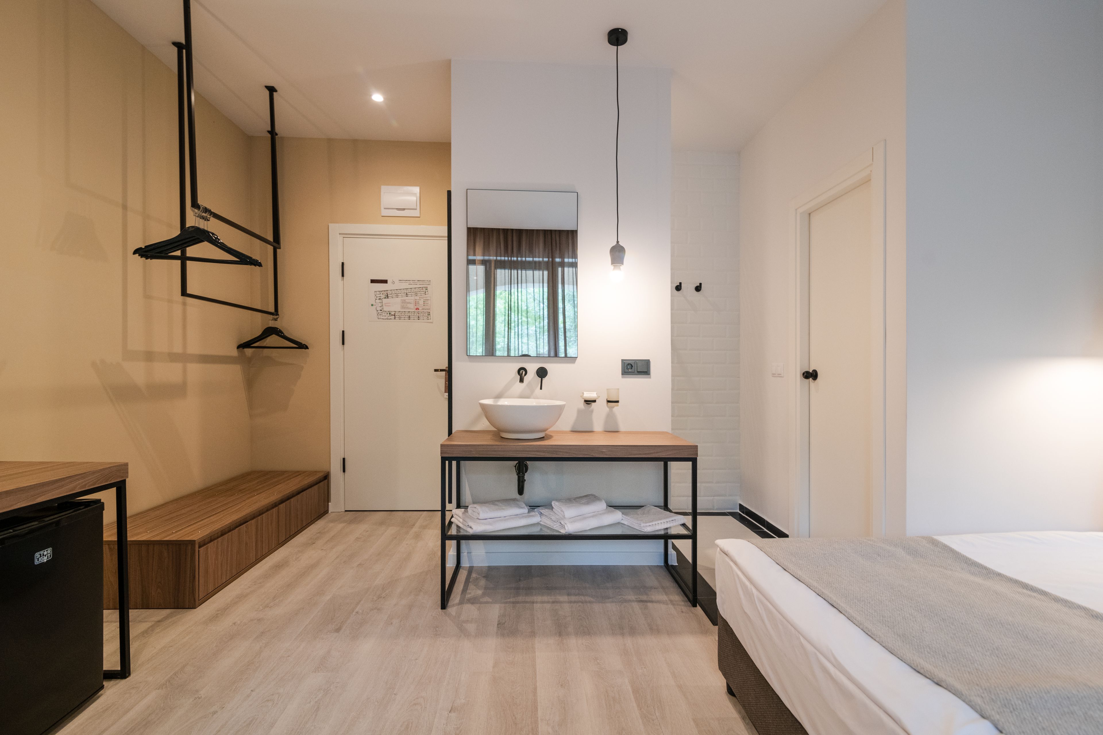 A modern hotel room with a wooden bench, a sink with a mirror, a bed, and a small table.
