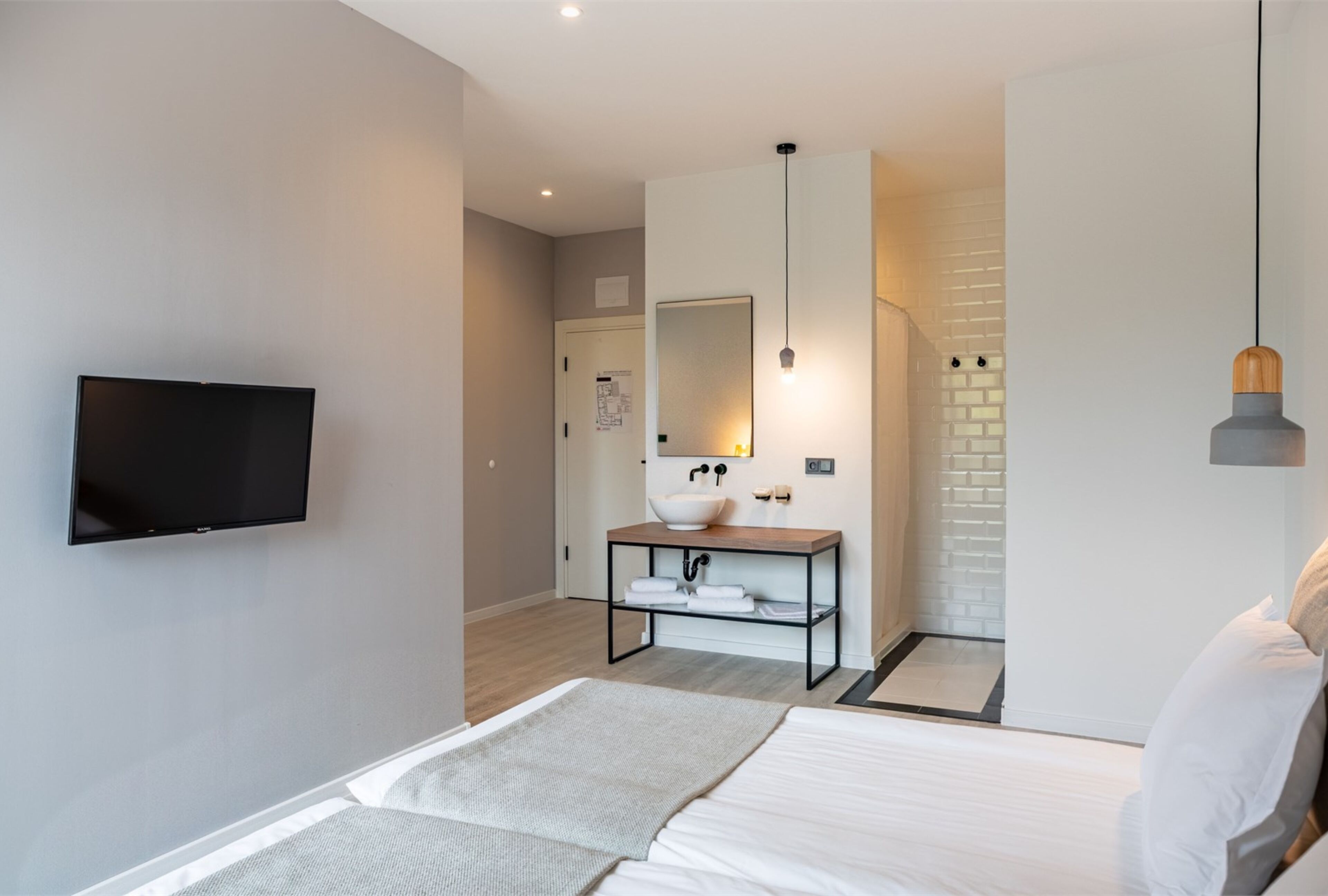 A modern hotel room with a flat-screen TV, a large mirror, a wooden bedside table, and a bed with white linens.
