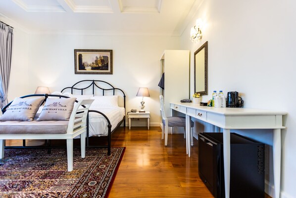 A hotel room with a bed, a desk, a chair, a small table, a mirror, and a refrigerator. - Merchant Villa (Chiang Mai)