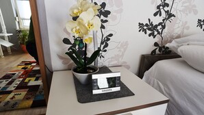 Une chambre d’hôtel bien rangée, avec un lit, une table de chevet sur laquelle se trouve un réveil, et une orchidée en pot.