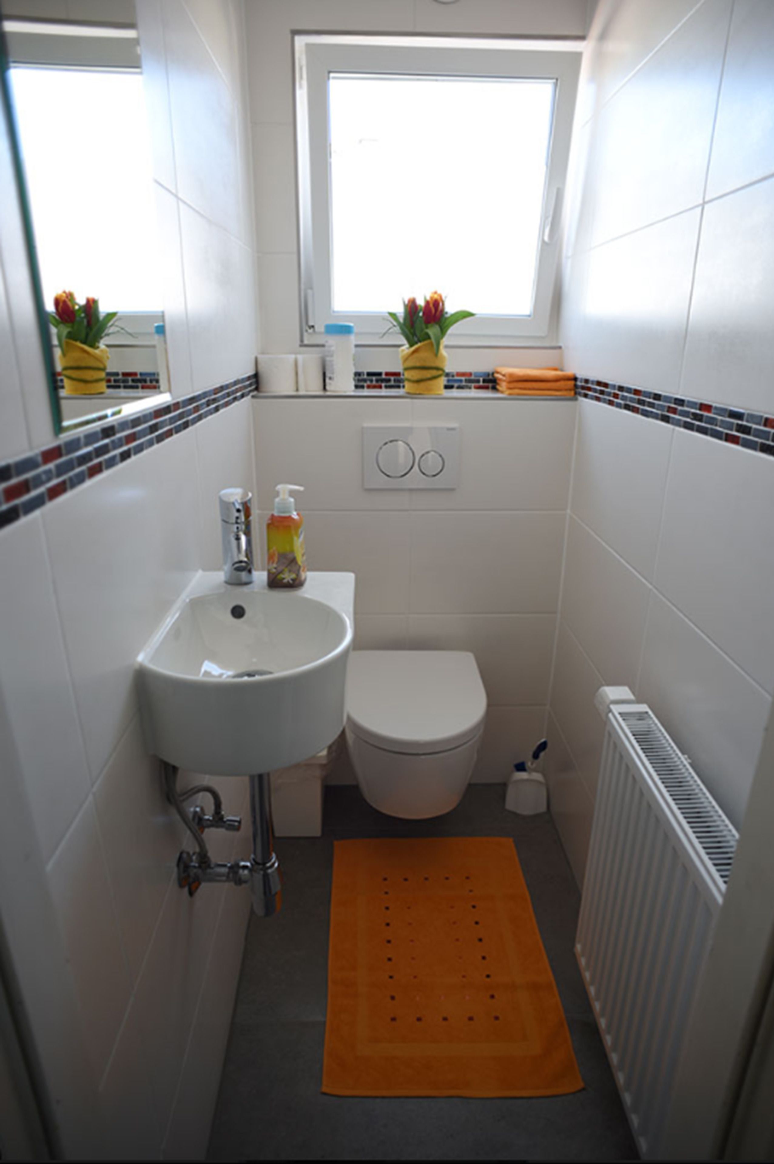 Une petite salle de bain propre, avec un lavabo, des toilettes et une fenêtre, le tout de couleur blanche. Une petite fenêtre se trouve au-dessus des toilettes, et on peut voir un porte-serviettes au mur.