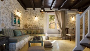 A rustic stone-walled living room with a wooden staircase, a sofa with patterned cushions, and a traditional fireplace. - Dotira, an Enchanting Heritage Home with Pool (Rethimno)
