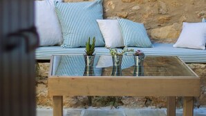 A stone wall with a rustic pot hanging, a cushioned bench with striped pillows, and a glass-top table with vases. - Dotira, an Enchanting Heritage Home with Pool (Rethimno)