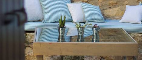A stone wall with a rustic pot hanging, a cushioned bench with striped pillows, and a glass-top table with vases.