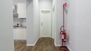 A narrow hallway with a white door at the end, a fire extinguisher mounted on the wall, and a kitchen area visible to the left. - Loft Rooms Trastevere (Rome)