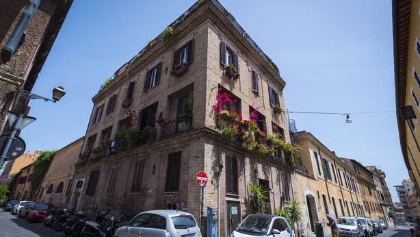A multi-story building with a brick facade, balconies with flowers, parked cars, and a clear blue sky. - Loft Rooms Trastevere (Rome)