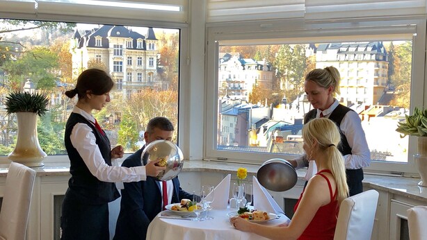 Spiseområde med et bord dækket til to, en tjener der serverer mad, og udsigt til bygninger gennem store vinduer.
