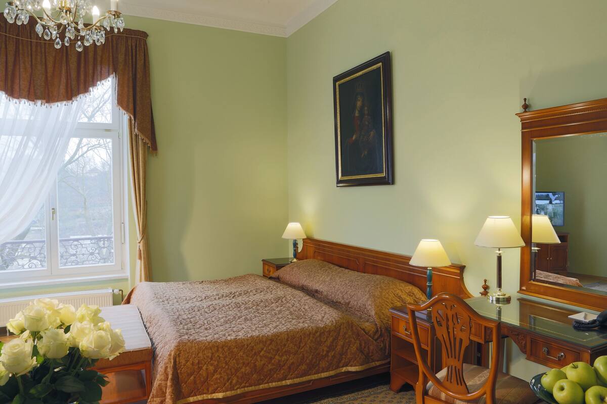 A neatly arranged hotel room with a bed, bedside lamps, a wooden chair, a desk with a mirror, and a vase of white flowers.