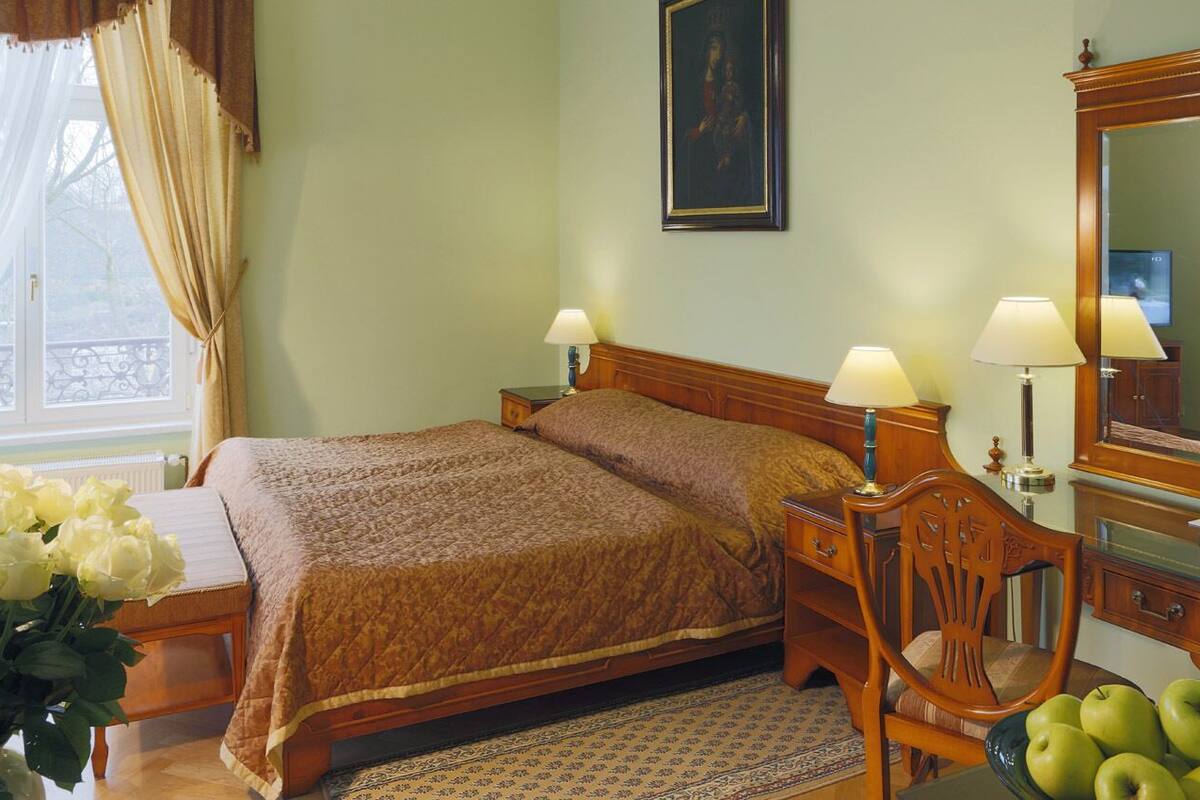 A hotel room with a bed, two bedside lamps, a wooden chair, a small table with a vase of flowers, and a mirror.