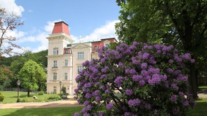 En stor, historisk bygning med rødt tag og mange vinduer, omgivet af frodig vegetation og lilla blomster.