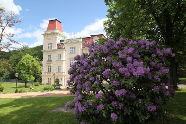 Um edifício grande e histórico com telhado vermelho e várias janelas, cercado por vegetação exuberante e flores roxas.