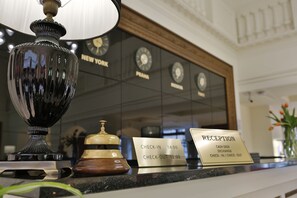 A reception desk with a check-in/out sign, a clock, and a decorative lamp. - Hotel Kolonada (Karlovy Vary)