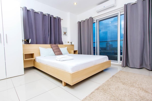 A modern bedroom with a large bed, grey curtains, a city view, and a white wardrobe.
