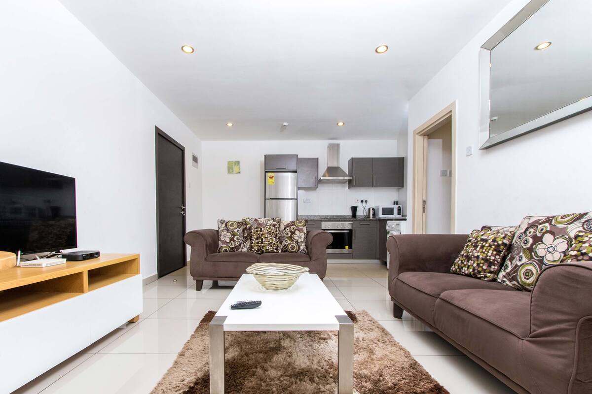 A modern living room with a flat-screen TV, two sofas, a coffee table, and a kitchen in the background.