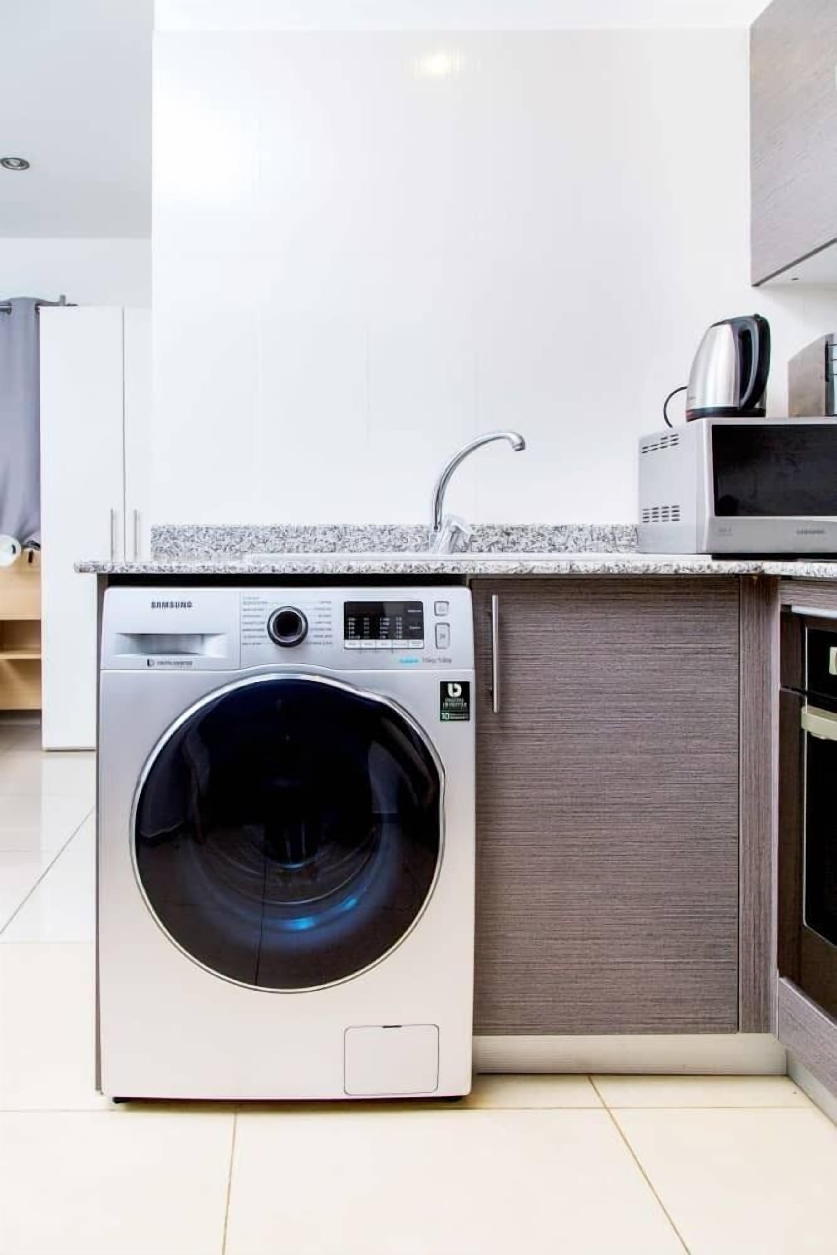 Una cocina compacta equipada con lavadora y secadora integradas, microondas y hervidor de acero inoxidable.