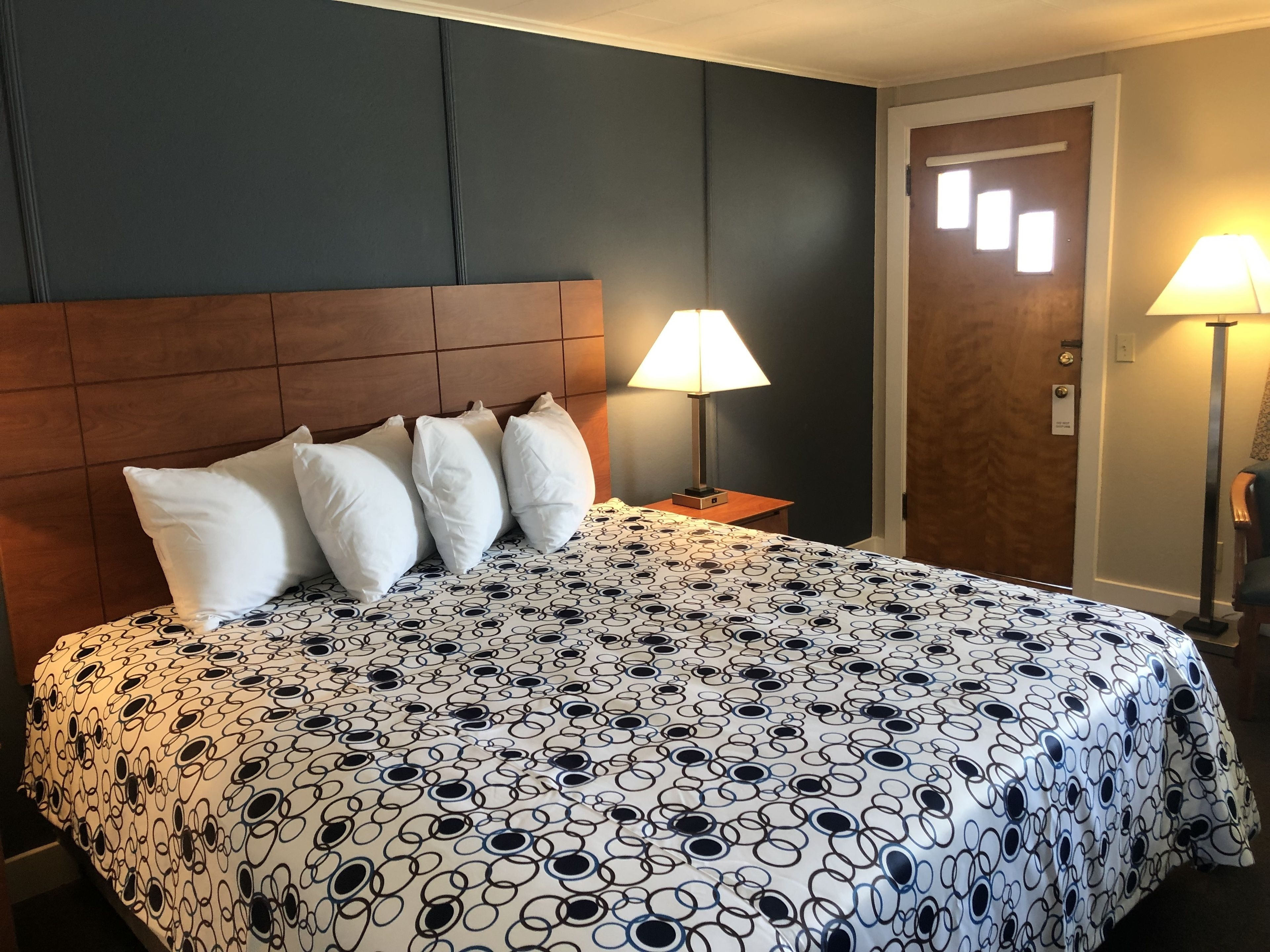 A hotel room with a large bed, a wooden headboard, white pillows, a patterned bedspread, two bedside lamps, and a door with a window.