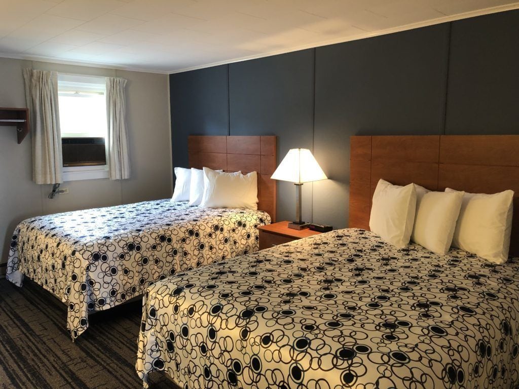 Une chambre d’hôtel avec deux lits, une table de chevet, une lampe, une fenêtre avec des rideaux et une étagère fixée au mur.