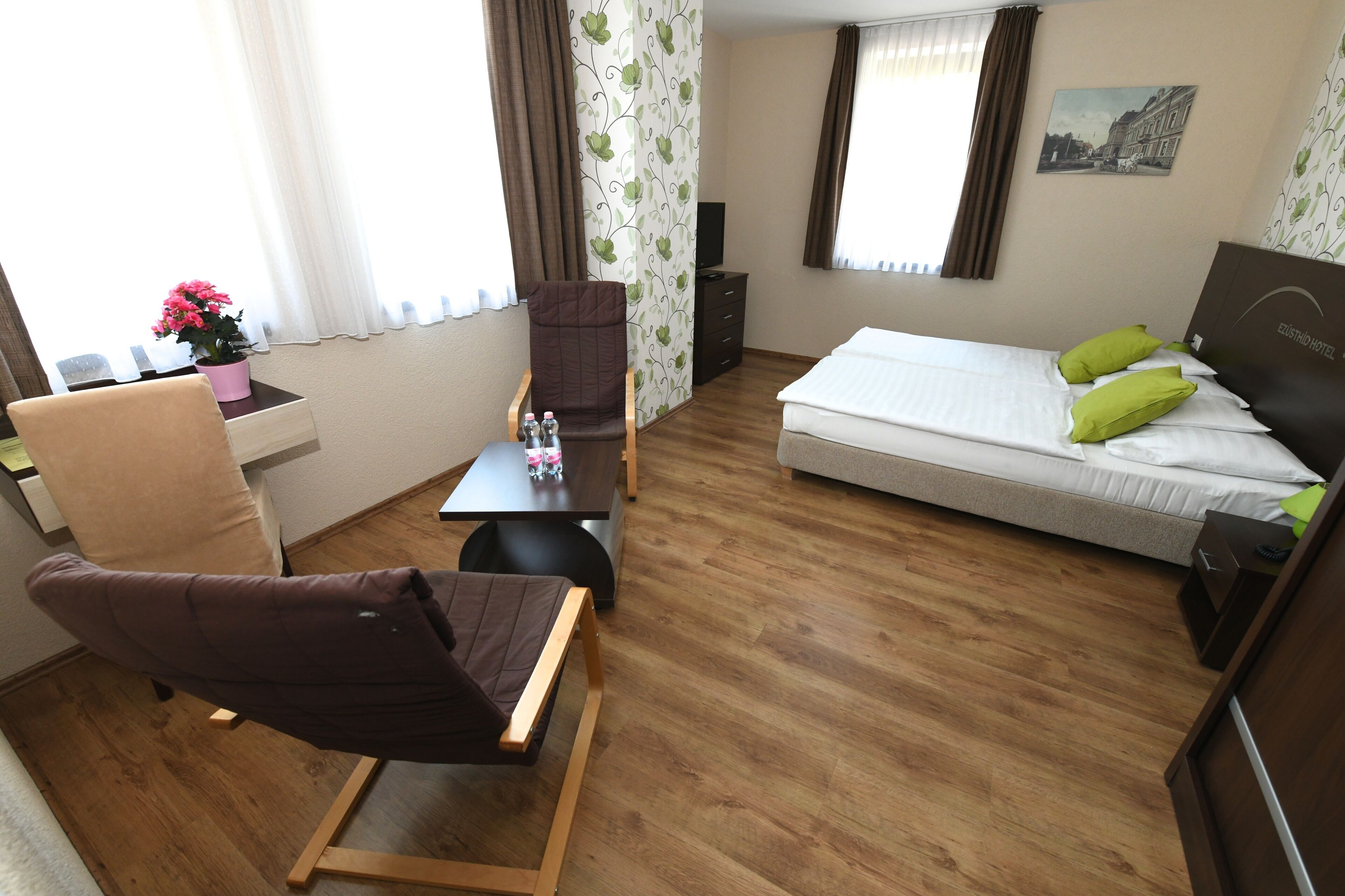 A hotel room with a bed, a chair, a small table, a desk, a potted plant, and a wall-mounted TV.