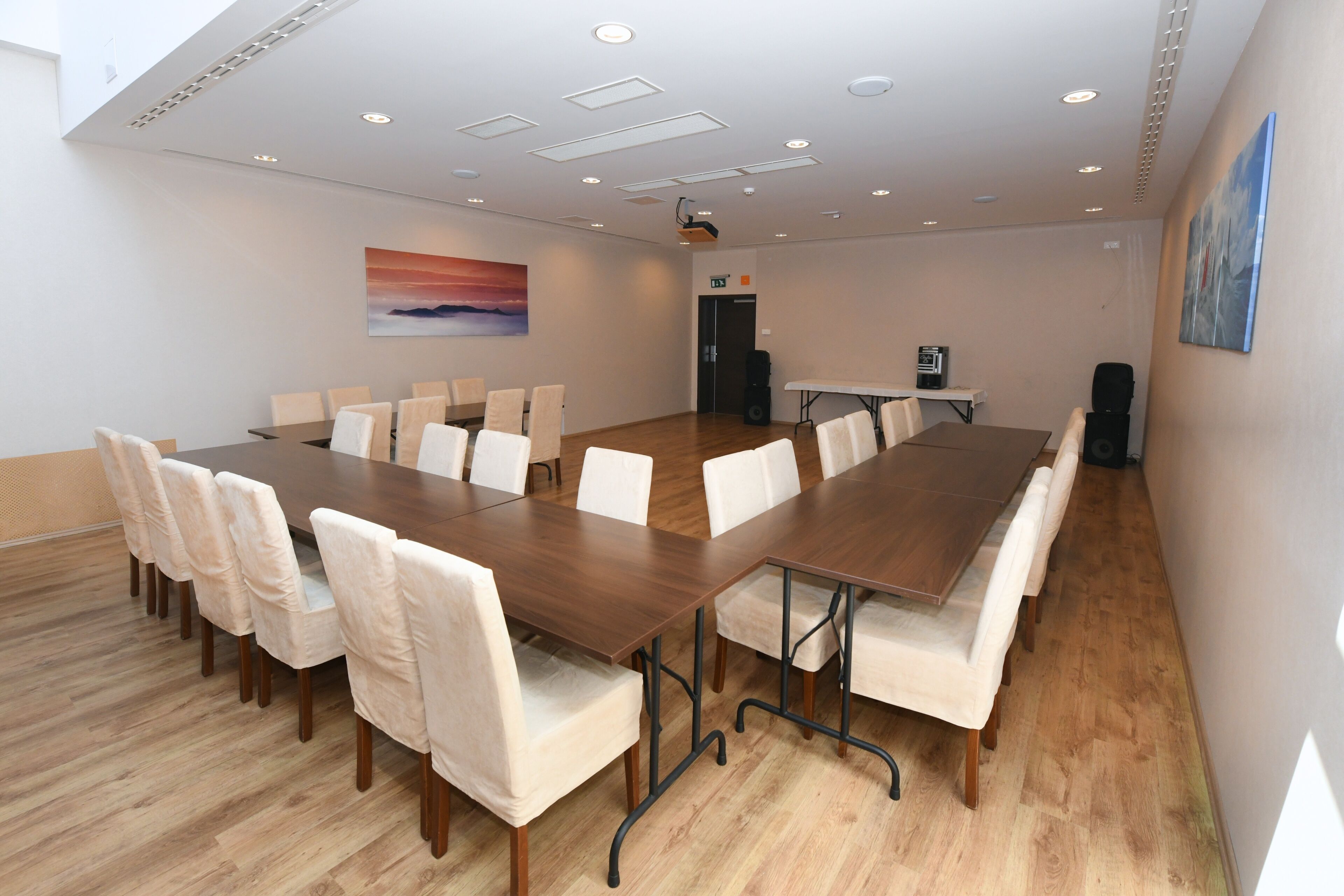 Un salón de conferencias con una larga mesa, sillas blancas, un proyector y un cuadro en la pared.