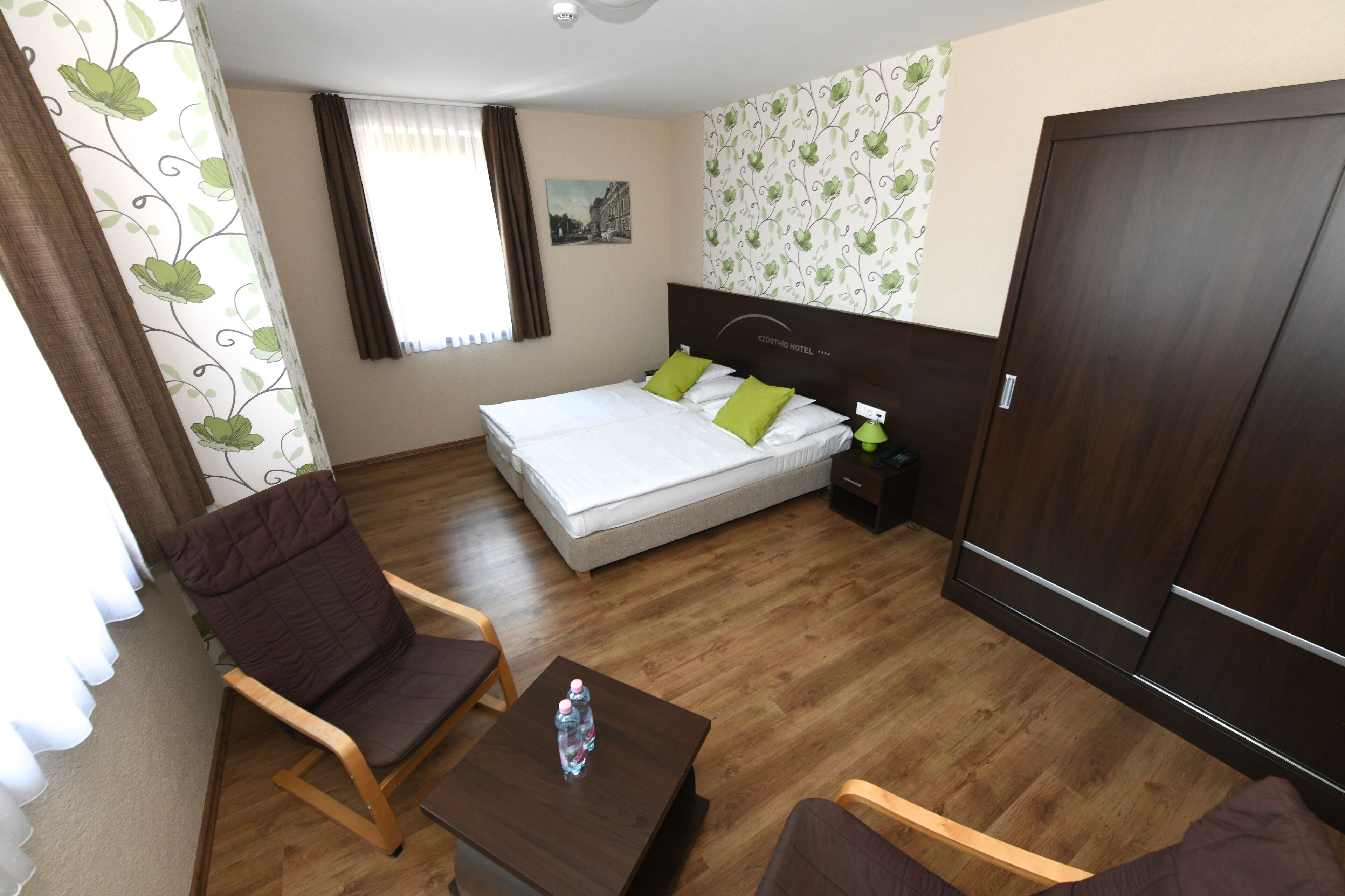 A hotel room with a bed, two chairs, a small table with water bottles, a nightstand, and a wardrobe.