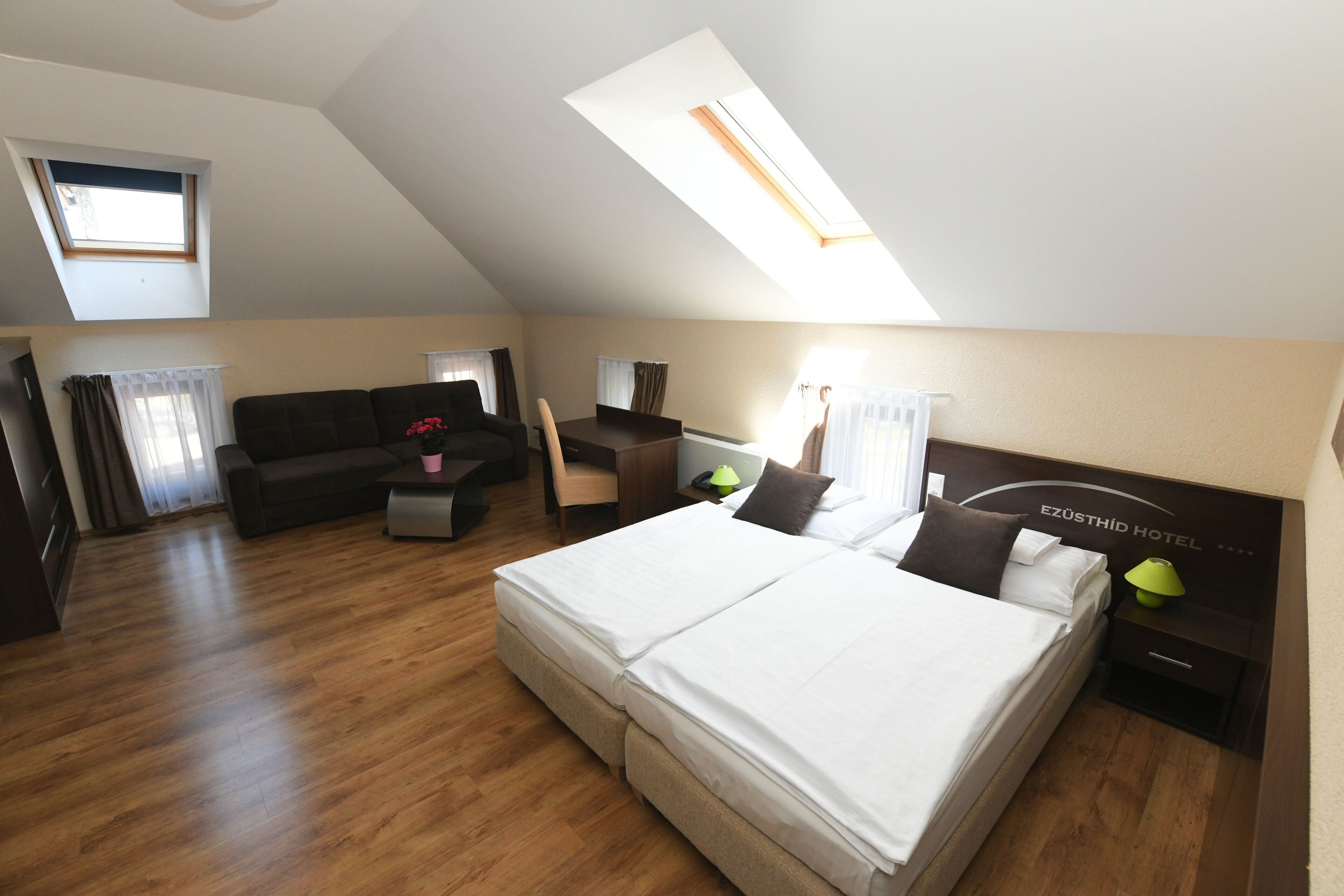A hotel room with a large bed, a desk, a chair, and a sofa. There is a skylight above the bed.