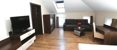 Un amplio loft con techo alto, tragaluz, un gran sofá oscuro, televisor de pantalla plana, escritorio de madera y zona de comedor.