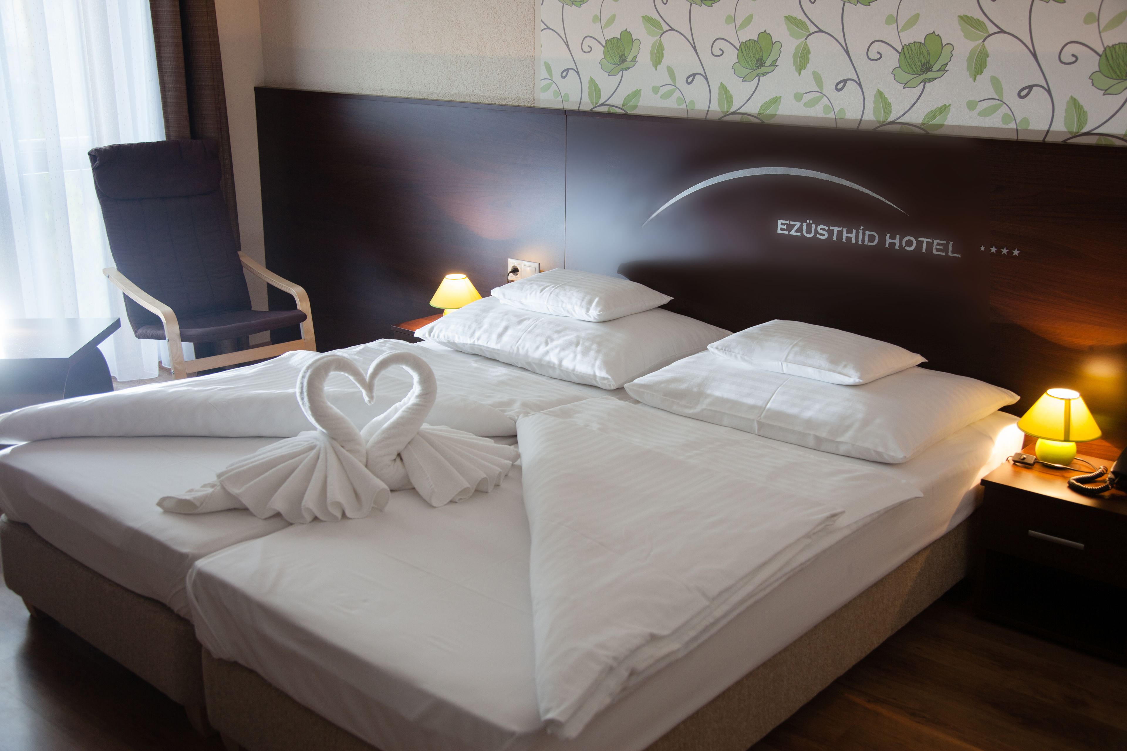 Una cama bien hecha con sábanas blancas y una disposición de toallas en forma de corazón, una silla y una mesita de noche con una lámpara.