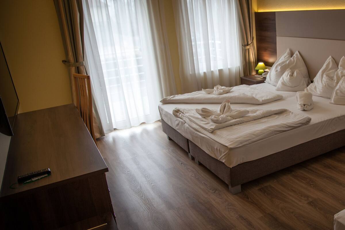 A hotel room with a bed, wooden floor, a nightstand with a lamp, and a TV on a wooden table.