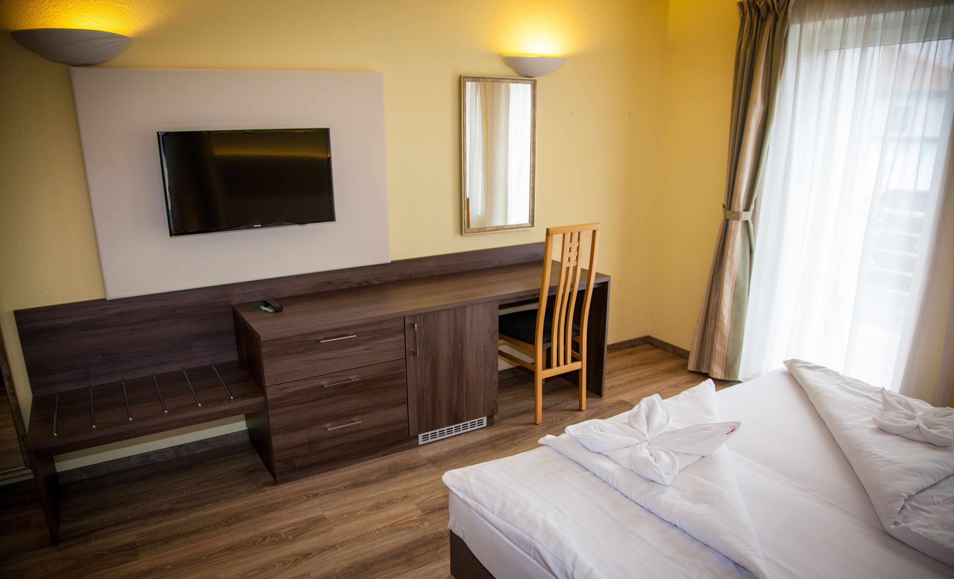 A hotel room with a bed, a desk with drawers, a television mounted on the wall, and a window with curtains.