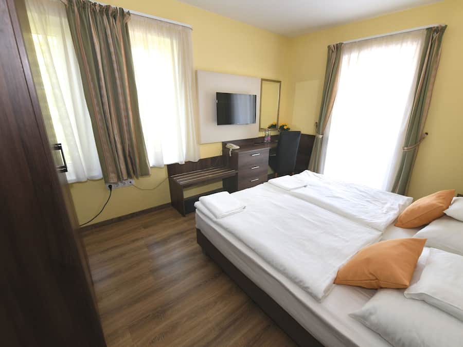 Quarto de hotel com cama, escrivaninha, cadeira, televisão e janela com cortinas.