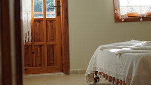 Ein Zimmer mit Holztür, einem Bett mit weißer Decke und einem Fenster mit Vorhang.