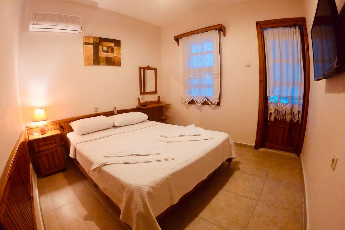 A neatly made bed with white linens and a bedside table with a lamp. A window with curtains is visible, and a flat-screen TV is mounted on the wall.