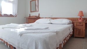 Ein ordentlich bezogenes Bett mit weißen Leinen und Spitzenbordüre, ein Nachttisch aus Holz mit einer Lampe und ein Fenster mit Vorhängen.