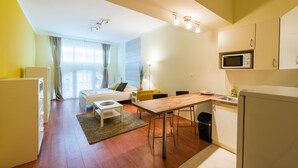 Un espacio habitable compacto con kitchenette, una cama con mesita de noche, una pequeña mesa de comedor y un ventanal con cortinas.