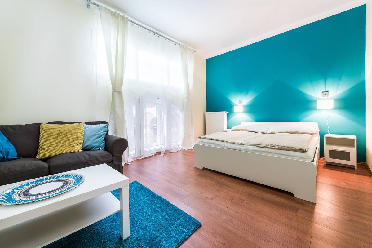 Un dormitorio moderno con cama, mesitas de noche, una mesita blanca, una alfombra azul, una mesa de centro blanca y un sofá con cojines.