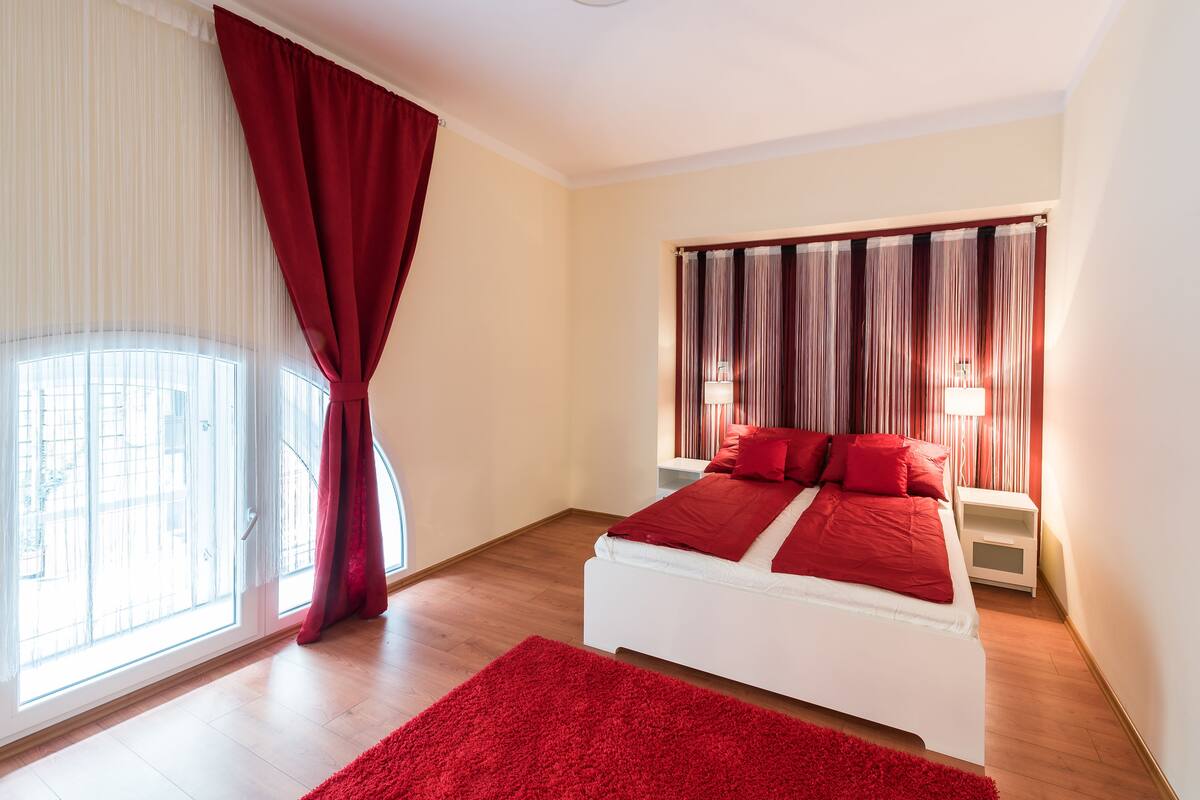 Un dormitorio con cama blanca, ropa de cama roja, alfombra roja, ventana con cortinas translúcidas y vista a edificios.