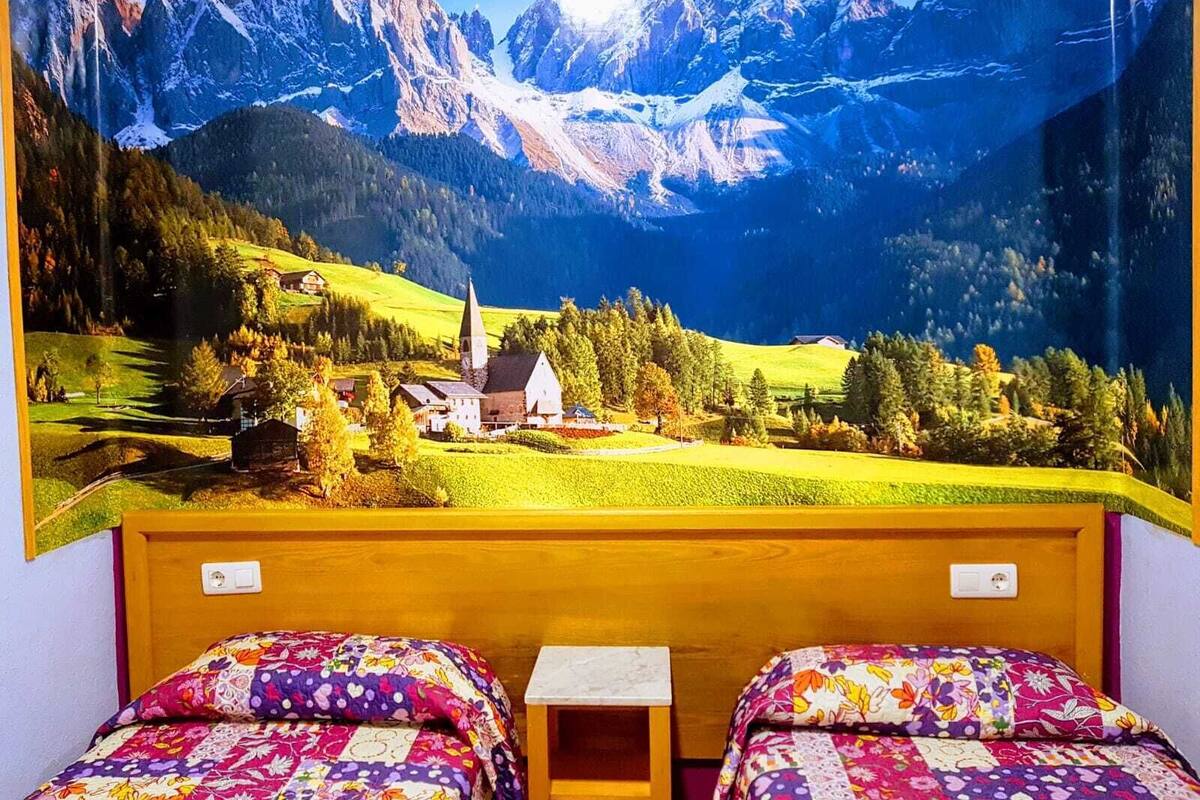 A room with two beds, a small table, and a mural of a mountainous landscape.