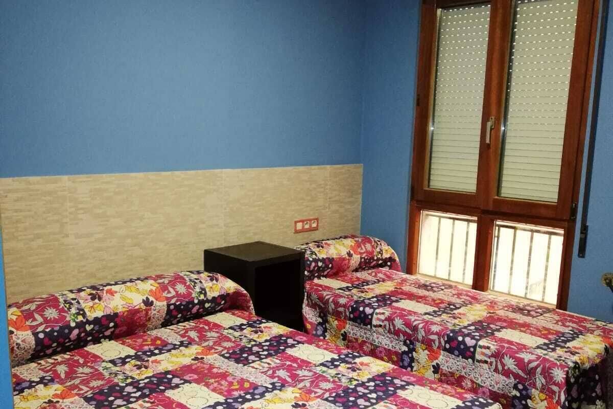 A room with two beds, a wooden headboard, a small table, and a window with blinds.
