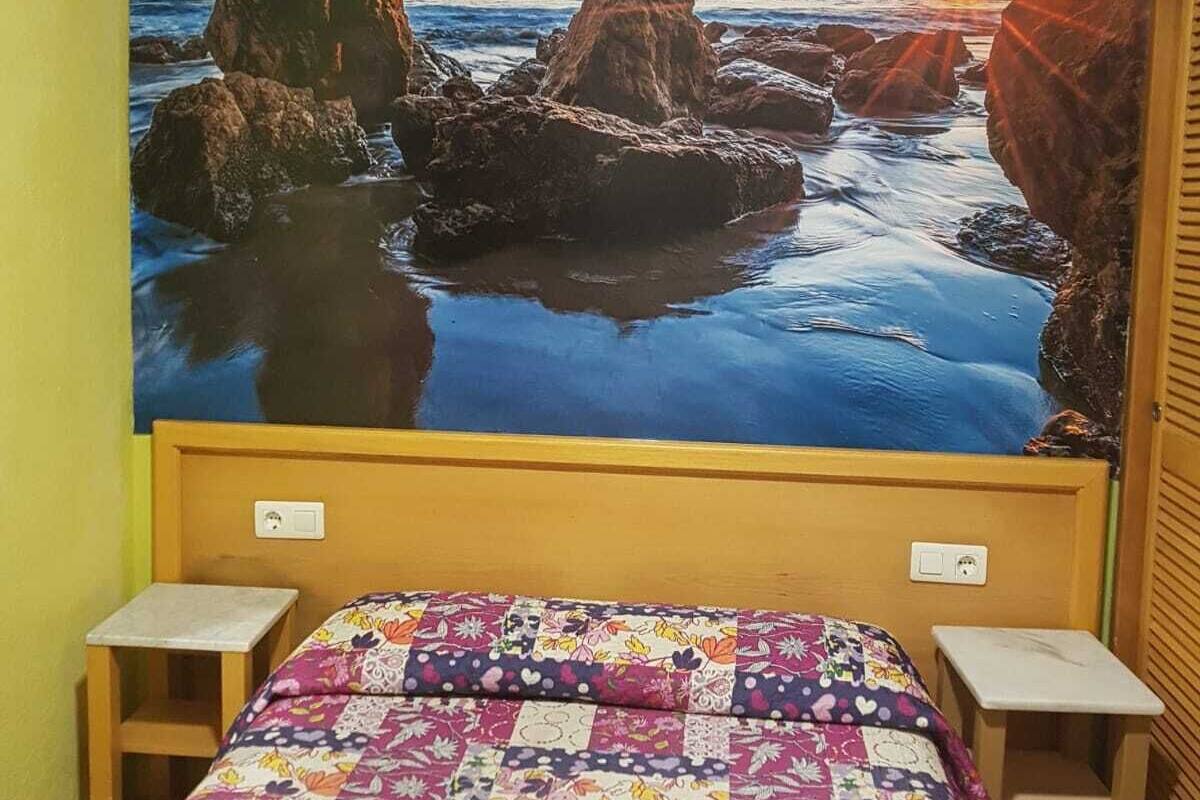 A bedroom with a bed, two bedside tables, and a large wall mural of a rocky coastline at sunset.