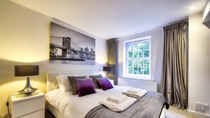 A neatly made bed with a cityscape painting above it, a bedside lamp, and a window with curtains.