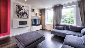 A modern living room with a grey sofa, a grey ottoman, a flat-screen TV, and a bull painting on the wall.
